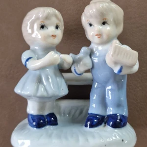 Två barn vid ett staket  - Detta är en porslinsfigur föreställande två barn vid ett staket, målad i ljusblå och vita toner. Figuren är i gott skick utan synliga sprickor eller större skador. Perfekt som prydnad eller samlarobjekt. Ingen märkning synlig på undersidan. Ca 9 cm lång och 10 cm hög.
