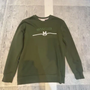 Grön sweatshirt från Moncler - Mörkgrön sweatshirt från Moncler med broderad logga och text på bröstet. Tröjan har rund halsringning, långa ärmar och ribbade muddar. Perfekt för dig som gillar stilrena och exklusiva plagg med en clean look.Och helt ny aldrig använd 