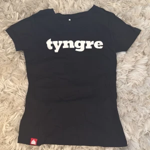 Svart t-shirt från Tyngre, S - Svart t-shirt från Tyngre i dammodell med vit logga tryckt över bröstet. Klassisk passform med rund hals och korta ärmar. Liten röd logolapp nertill. Perfekt för dig som gillar enkel och sportig stil. Aldrig använd