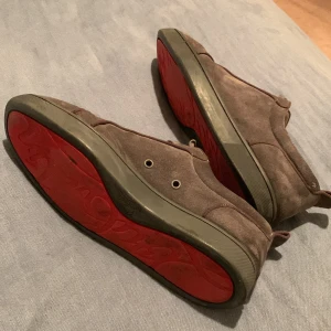 Gråa mocka skor från Christian louboutin  - Snygga bruna sneakers i mocka med klassisk snörning och ikonisk röd sula. Skorna har rund tå och diskreta detaljer, samt beige insida i skinn. Perfekta för dig som vill sticka ut med en stilren men unik look.