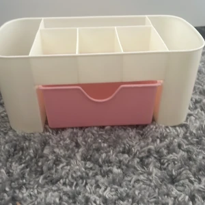 Plastförvaringslåda med fack och utdragbar låda - Praktisk plastförvaringslåda med flera fack upptill och en utdragbar rosa låda. Perfekt för att organisera småelektronik, tillbehör eller skrivbordstillbehör. Produkten är i gott skick utan synliga skador eller slitage.