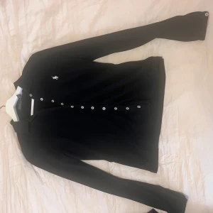 Svart pikétröja från Polo Ralph Lauren - Svart långärmad pikétröja från Polo Ralph Lauren med klassisk krage och vita knappar hela vägen framtill. Tröjan har det ikoniska broderade logotypen i vitt på bröstet och är gjord i mjuk bomull.  Storlek S, passar nog även M 🖤