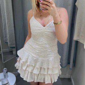 Vit kort volangklänning med guldtrådar - Supersöt vit kort klänning med tunna axelband, v-ringning och flera lager volanger nertill. Klänningen har rynkade detaljer framtill och är dekorerad med diskreta guldtrådar. Helt ny och aldrig använd🩷