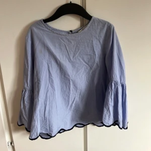 Blåvit randig blus från Zara  - Söt blus från Zara i blått och vitt med tunna ränder. Vida, trumpetformade ärmar och svarta vågiga kanter nertill och vid ärmslut. Tillverkad i bomull och har en liten knapp i nacken. Perfekt för dig som gillar en luftig och trendig look.