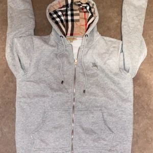 Grå hoodie från Burberry L/XL - Säljer en grå hoodie från Burberry i storlek XL. Klassisk dragkedja framtill, huva med ikoniskt rutigt foder och snörning. Broderad logga på bröstet och två stora fickor. Perfekt för dig som vill ha en stilren och lyxig vardagslook. Påsse ingår med hoodien