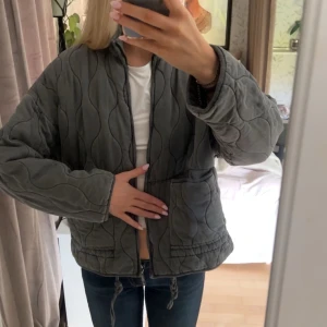 Grå quiltad jacka från Zara - Quiltad grå jacka från Zara med vågigt mönster, två stora fickor framtill och dragsko i nederkant. Jackan har en avslappnad passform och stängs med dragkedja. Perfekt för lager-på-lager och snygg till jeans.