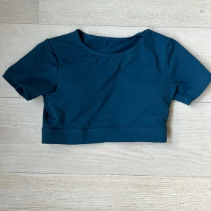 NVGTN träningstopp - Säljer en mörkblå croppad träningstopp från NVGTN med korta ärmar och rund hals. Toppen har en öppen rygg med vit insida och inbyggda kupor för extra stöd. Perfekt för gymmet eller andra sportiga aktiviteter.