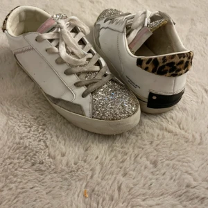 Glittriga sneakers från Crime - Snygga vita sneakers från Crime med glittriga silverdetaljer på tån och plösen. Baktill har de en cool leopardmönstrad häl och svarta detaljer. Skorna har snörning och en klassisk rund tå. Perfekta för dig som vill sticka ut med din stil.