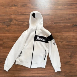 Vit Moncler hoodie med svart detalj - Snygg vit hoodie från Moncler med svart panel och broderad logga på ärmen. Dragkedja framtill, huva och två fickor. Stora Moncler-bokstäver på bröstet och svart detalj över ena ärmen ger en cool kontrast. Perfekt för dig som gillar streetwear.