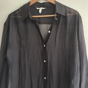Svart transparent skjorta från H&M - Svart transparent skjorta från H&M med klassisk krage och vita knappar framtill. Skjortan har lång ärm och är tillverkad i ett tunt, lätt och flowy material som ger en cool och edgy vibe. Perfekt att styla lager på lager eller över ett linne.