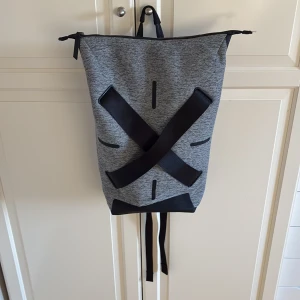 Grå och svart ryggsäck med laptopfack från Li-Ning - Snygg ryggsäck i gråmelerat tyg med svarta detaljer och breda svarta remmar i kors på framsidan. Väskan har vadderade axelband, dragkedja upptill och ett separat svart laptopfack med dragkedja och pennhållare. Från Li-Ning. I fint skick.