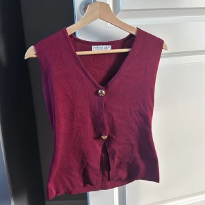 Vinröd stickad väst från Design by Amor - Snygg vinröd stickad väst med v-ringning och två dekorativa knappar framtill. Perfekt att styla över en t-shirt eller skjorta för en cool layering-effekt. Västen har en klassisk passform och är gjord i mjukt stickat material.