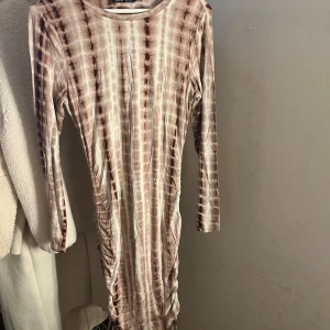 Mönstrig  långklänning från SHEIN - Säljer en långärmad långklänning från SHEIN i storlek M. Klänningen har ett beige och brunt randigt mönster, rund halsringning och snygga rynkade detaljer längs sidorna. Materialet är mjukt och stretchigt, perfekt för dig som gillar en bekväm men ändå utstickande look.