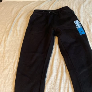 Svarta mjukisbyxor från Hoodrich XL - Svarta mjukisbyxor från Hoodrich i storlek XL för ungdomar. Byxorna har resår och snörning i midjan samt broderad HOODRICH-logga i vitt och blått på ena benet. Tillverkade i mjukt material för maximal komfort och avslappnad stil.