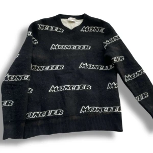 Svart Moncler stickad tröja  - Svart stickad tröja från Moncler med vit logotyp tryckt över hela plagget. Tröjan har rund halsringning och långa ärmar. Perfekt för dig som gillar streetstyle och vill sticka ut med en exklusiv look.