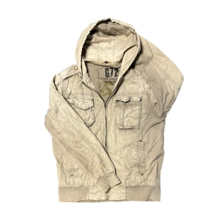 Beige field jacka med huva från G72 - Snygg beige fieldjacka från G72 med huva och dragkedja framtill.  Vintage utseende med en snygg fade och lite wear. Finns en liten fläck på ryggen (se bild 5), men tror att det går bort lätt då jag inte har försökt.