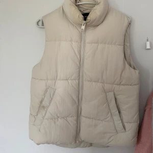 Beige dunväst från Lindex - Säljer en beige dunväst från Lindex med hög krage och dragkedja framtill. Västen har två snedställda fickor och quiltad design som ger en puffig look. Perfekt att slänga över en hoodie för en chill stil. Lite missfärgad vid kragen lite osäker om det går bort eller inte. Dock inget man tänker på när den sitter på.