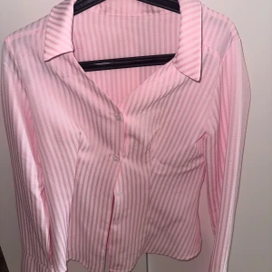 Rosa randig skjorta med slits - Snygg rosa skjorta med vita ränder och klassisk krage. Skjortan har lång ärm, knappar framtill och en cool slitsdetalj i sidan. Perfekt för dig som gillar en fräsch och trendig look. Materialet känns lätt och luftigt. Använd 2 gånger.