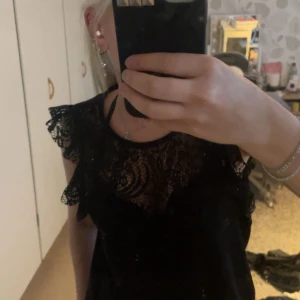 Svart spetsklänning från Vero Moda - Supersnygg svart klänning från Vero Moda i storlek S. Klänningen har vackra spetsdetaljer upptill, volang vid axlarna och är ärmlös. Perfekt för dig som vill ha en stilren men ändå detaljrik look. Stängs med knapp i nacken.