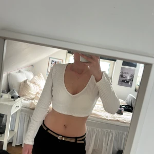 Vit långärmad topp - Säljer en vit croppad topp med lång ärm och djup v-ringning. Toppen är ribbad och sitter tight, perfekt för dig som gillar en enkel men snygg stil. Materialet känns mjukt och stretchigt, passar till många olika outfits.