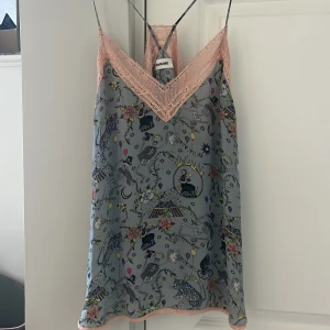 Mönstrad linne från Zadig & Voltaire - Supersöt linne med tunna axelband från Zadig & Voltaire. Grå botten med färgglatt, lekfullt mönster av skelett, leoparder, blommor och ballonger. V-ringad med ljusrosa spetsdetaljer vid hals och nederkant. Perfekt för dig som gillar unika prints. 