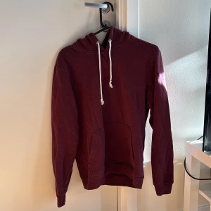 Vinröd hoodie med snörning - Vinröd hoodie med vit snörning i huvan och stor magficka framtill. Tröjan har lång ärm och en klassisk loose passform, perfekt för chill dagar. Materialet är mjukt och känns skönt mot huden.