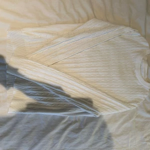 Vit kabelstickad tröja med rund hals - Snygg vit kabelstickad tröja med rundad hals och ribbade muddar vid ärmslut och nederkant. Tröjan har ett klassiskt mönster med diskreta tvinnade ränder och är långärmad. Perfekt för dig som gillar stilrena och tidlösa plagg.