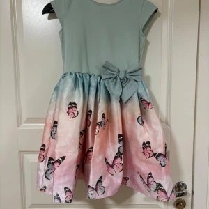 Mintgrön klänning med fjärilar H&M - Superfin mintgrön klänning från H&M med korta ärmar, stor rosett i midjan och kjol i pastellfärger med fjärilsmönster i rosa och blått. Kjolen har en lätt glansig finish och dragkedja bak. Perfekt för dig som gillar färg och detaljer!