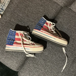 Höga sneakers med USA-flagga och nitar - Säljer ett par höga sneakers med amerikansk flaggdesign i rött, vitt och blått. Skorna har vita snören, rund tå och platt sula. Baksidan är dekorerad med silvriga stjärnnitar på blå bakgrund. Materialet är canvas och detaljer i syntet. Använda endast ett fåtal gånger. Står i skon att det är från Duffy.