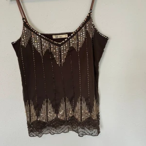 Brun paljettlinnetopp från Flash Basic - Säljer en brun linne-topp med tunna axelband från Flash Basic. Toppen har glittriga paljettdetaljer i guld och silver framtill, samt ett geometriskt mönster längs halsringningen och nederkanten. Materialet är lätt och syntetiskt, perfekt för att sticka ut.
