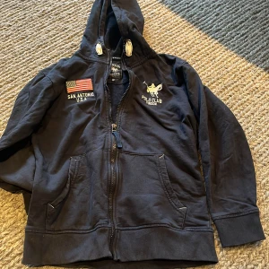 Svart hoodie Polo Club Vinson - Svart hoodie från Polo Club Vinson med broderad logga och amerikansk flagga på bröstet. Tröjan har huva med snörning, två fickor fram och dragkedja. Materialet är mjuk bomull och passformen är normal. Perfekt för dig som gillar sportig och avslappnad stil.