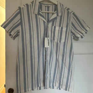 Randig kortärmad skjorta från Kappahl - Fräsch skjorta från Kappahl i storlek 158 med vertikala blå och vita ränder. Skjortan har korta ärmar, klassisk krage, bröstficka och knäppning framtill. Tillverkad i lätt och skönt bomullsmaterial, perfekt för varma dagar.