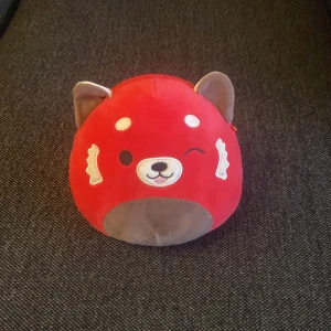Röd Panda - Bilderna visar ett rött gosedjur som liknar en räv eller panda, med broderat ansikte och mjuka öron. Produkten ser ut att vara i mycket gott skick utan synligt slitage. Perfekt som present eller mysig inredningsdetalj.