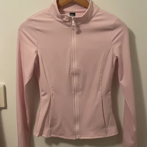Ljusrosa vindjacka från Dazy - Snygg ljusrosa vindjacka från Dazy i storlek M. Jackan har en smickrande slim passform, hög krage och hel dragkedja framtill. Tillverkad i mjukt syntetmaterial som känns skönt mot huden. Perfekt för dig som gillar sportig och clean stil.