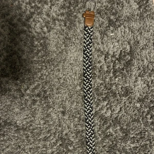 Svartvit flätad bälte med skinn - Snyggt flätat bälte i svart och vitt med bruna skinn-detaljer vid spännet och änden. Bältet har ett coolt zigzag-mönster och är perfekt för att ge outfiten lite edge. Passar till jeans eller chinos och har en klassisk metallspänne.