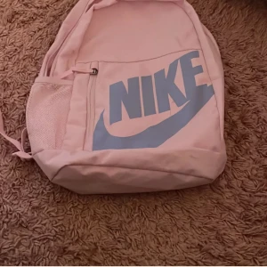 Ljusrosa Nike ryggsäck med blå logga - Säljer en ljusrosa ryggsäck från Nike med stor blå logga framtill. Väskan har ett stort huvudfack, ytterficka med dragkedja och meshficka på sidan. Justerbara, vadderade axelremmar för extra komfort. Perfekt för skolan eller träningen.