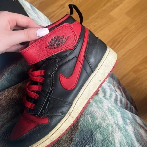 Nike Air Jordan 1 svart/röd high - Säljer ett par Nike Air Jordan 1 high sneakers i svart och rött skinn med klassisk röd swoosh och Jordan Wings-logga på sidan. Skorna har snörning, dragkedja på sidan och rund tå. Sulan är vit med röd yttersula. Perfekt för dig som gillar retro och streetstyle.