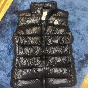 Svart dunväst från Moncler - Snygg svart dunväst från Moncler med glansig finish och klassisk logga på bröstet. Västen har quiltad design, hög krage och dragkedja framtill. Perfekt för lager-på-lager och ger en cool vibe till din outfit. (DET FINNS NFC)