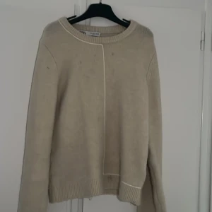 Beige stickad tröja från Zara (S) - Mysig beige stickad tröja från Zara med rund halsringning och diskret sömdetalj framtill. Tröjan har långa ärmar och ribbade muddar vid hals, ärmslut och nederkant. Perfekt för lager-på-lager under kyliga dagar.