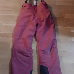 Rosa täckbyxor med svarta detaljer - Säljer ett par rosa täckbyxor med svarta förstärkningar vid bensluten och justerbara hängslen. Perfekta för vinteraktiviteter och håller dig varm och torr. Byxorna har elastisk midja och är tillverkade i slitstarkt syntetmaterial. 