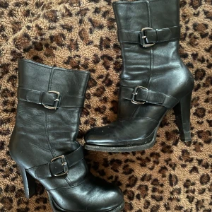 Svarta boots med spännen och klack - Snygga svarta boots i skinn med höga klackar och dekorativa spännen runt skaftet och vristen. De har rund tå och en stilren design som passar perfekt till både jeans och kjol. Ger en edgy vibe till din outfit.