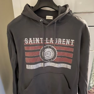 Grå Saint Laurent hoodie med tryck - Säljer en grå hoodie från Saint Laurent med vintageinspirerat tryck i rött och vitt framtill. Tröjan har huva med snörning och en stor magficka. Perfekt för dig som gillar streetstyle och exklusiva märken. Icke orginal toppkvalitet