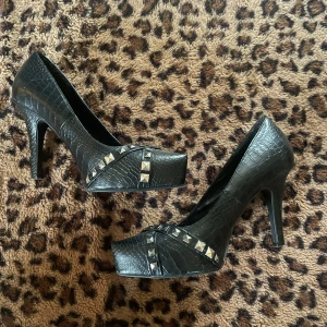 Svarta högklackade skor med nitar från Din Sko - Snygga svarta högklackade skor från Din Sko med krokopräglat mönster och silverfärgade nitar. Skorna har spetsig tå, hög klack och plattform framtill. Tillverkade i syntetmaterial med cool rockig vibe. Perfekta för dig som vill sticka ut.