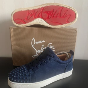 Blåa Christian Louboutins - Säljer ett par blå sneakers från Christian Louboutin i mocka med ikoniska röda sulor och coola nitar framtill. Skorna har snörning och vit platt sula, samt lyxiga detaljer som gör dem till ett riktigt statement. Perfekta för dig som vill sticka ut med exklusiv stil. Priset kan diskuteras. Rätt så nya. Bara skriva vid någon fråga!