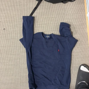 Mörkblå sweatshirt från Polo Ralph Lauren - Säljer en mörkblå sweatshirt från Polo Ralph Lauren med klassisk rund hals och röd broderad logga på bröstet. Tröjan har långa ärmar och är tillverkad i mjuk bomull, perfekt för en chill och stilren look.