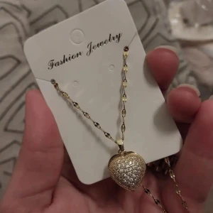 Snygg halsband med hjärtberlock i rostfritt stål med 18k guldpläterad  - Snyggt halsband i rostfritt stål med 18k guldpläterad med en hjärtformad berlock som öppnas och har en vit pärla inuti. Berlocken är glittrig med små stenar på framsidan och kedjan har dekorativa länkar. Perfekt för dig som gillar detaljer och hjärtan.