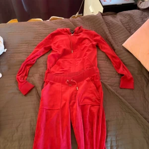 Röd Juicy Couture hoodie i velour - Juicy Couture hoodie i röd velour med huva och dragkedja framtill. Klassisk sportig look med långa ärmar, ribbade muddar och fickor framtill. Perfekt för en avslappnad och trendig stil.