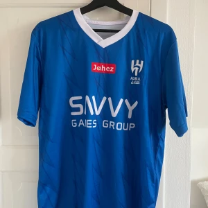 Al Hilal Neymar JR blå fotbollströja - Blå fotbollströja från Al Hilal med Neymar JR och nummer 10 på ryggen. Tröjan har vit V-ringad krage, korta ärmar och flera sponsorer tryckta fram och bak. Materialet är lätt och andas, perfekt för match eller träning. Snygga subtila mönster i tyget ger extra stil.