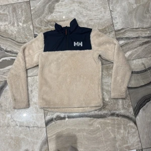 Beige fleece-tröja från Helly Hansen - Mysig beige fleece-tröja från Helly Hansen med marinblått parti upptill och HH-logga på bröstet. Tröjan har half zip med orange detalj, hög krage och långärmade ärmar. Perfekt för kalla dagar när du vill vara både chill och varm.