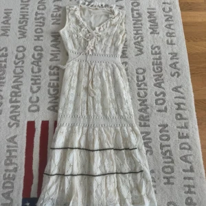 Vit långklänning med spets och snörning - Superfin vit långklänning med spetsdetaljer och blommigt mönster. Klänningen har snörning vid halsen, markerad midja och är ärmlös. Materialet är bomull med spets och broderade blommor, perfekt för sommarens alla tillfällen. Romantisk och bohemisk vibe!